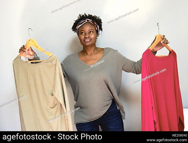 Portrait of young black woman with afro hairstyle with clothes on hangers indoor at home. Lifestyle concept