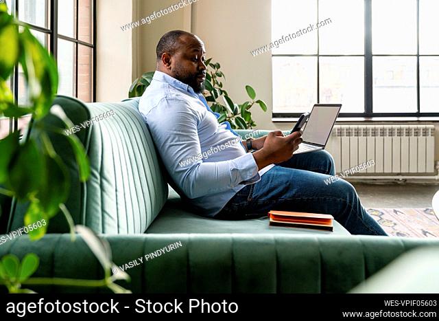 Businessman holding laptop using mobile phone sitting on sofa in office