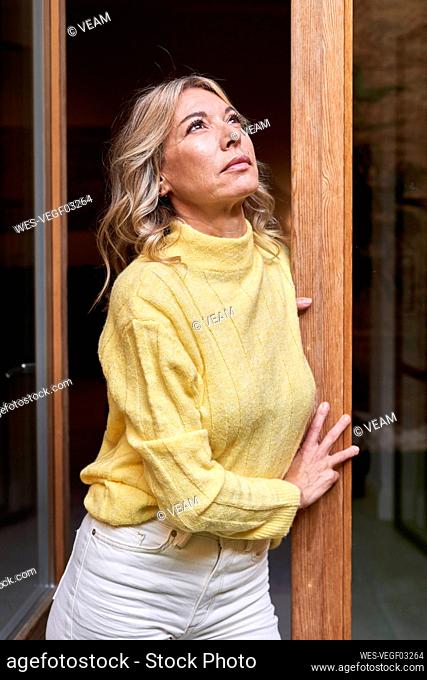 Thoughtful mature woman in yellow sweater looking up while leaning on door at home