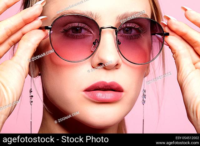 Close-up portrait of attractive young woman in tinted glasses. Female looking at camera on pink background