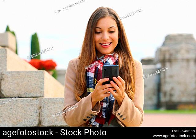 Pretty cheerful young woman messaging with mobile phone sitting on stairs in winter time
