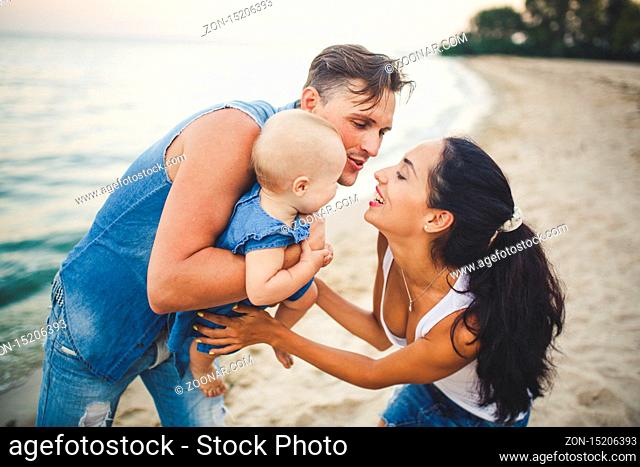 A young, beautiful family of three. Mom, Dad and daughter in the arms of my father play, rejoice, smile on the sandy beach on the beach in the summer