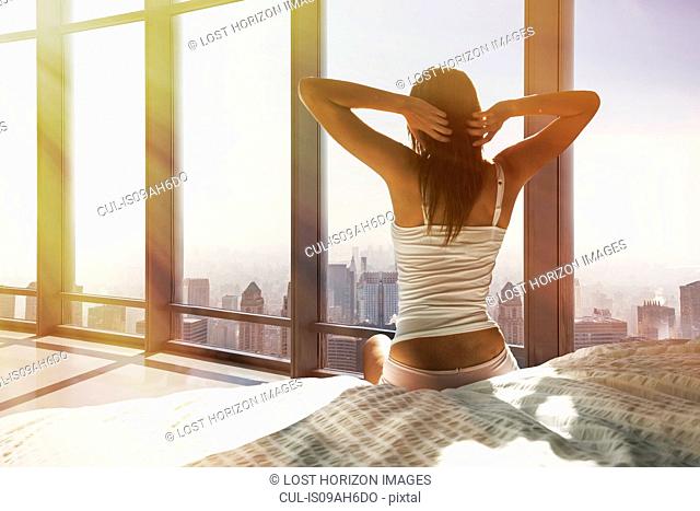 Young woman sitting on bed, stretching, overlooking city