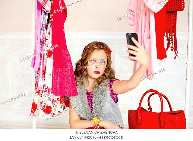 the little girl with red lipstick on lips tries on clothes of red color and looks in phone. She sits on a mobile hanger, and nearby there is a red leather bag