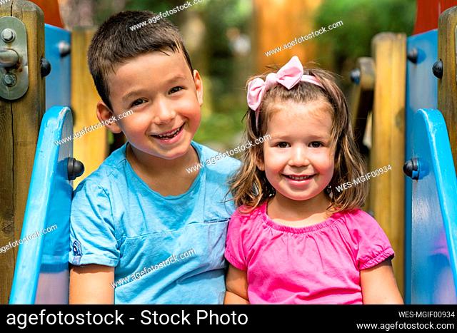 Portrait of happy little boy and his younger sister sitting together on playground slide