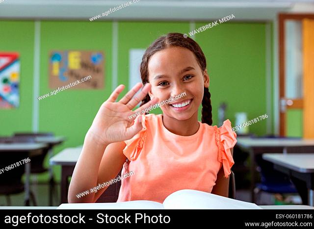 Portrait of smiling mixed race schoolgirl sitting at desk in classroom waving hand