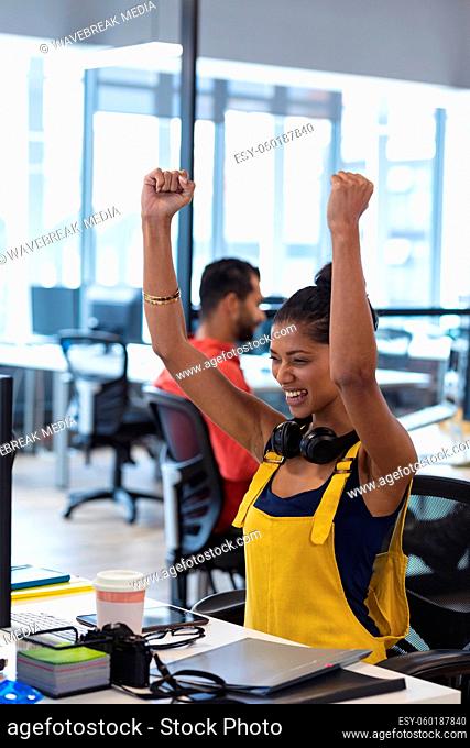 Mixed race female creative sitting at desk celebrating raising her arms and smiling