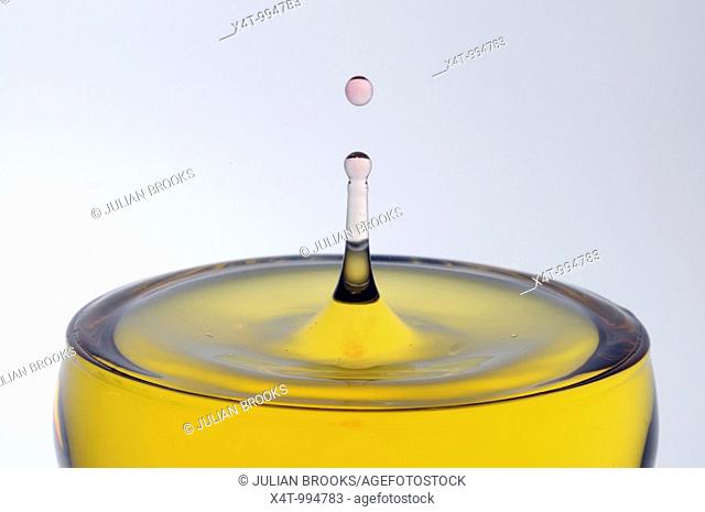 A drop of pink water falling into a glass of yellow liquid forming a column with a drop on top