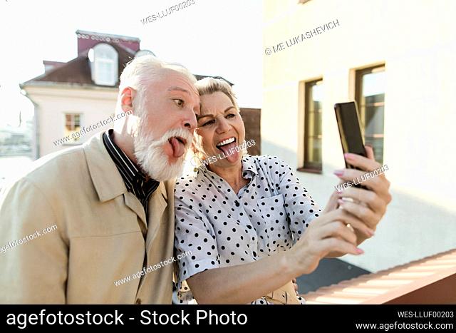 Couple sticking out tongue while taking selfie through smart phone on rooftop