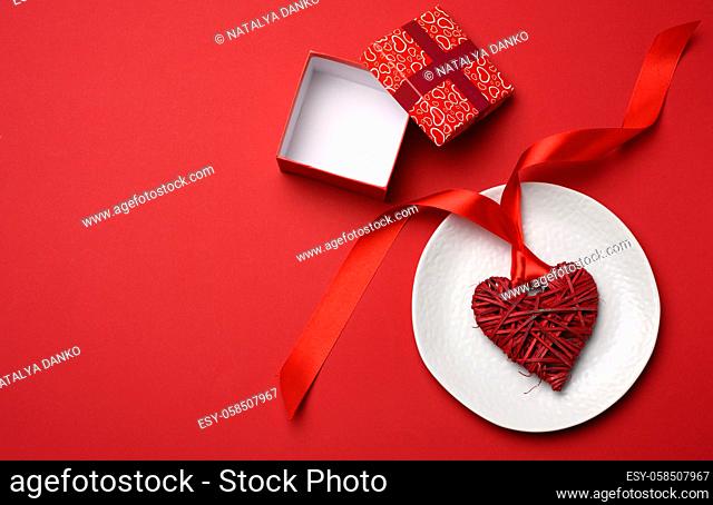 open square cardboard box and wicker heart with silk ribbon in white ceramic plate, top view