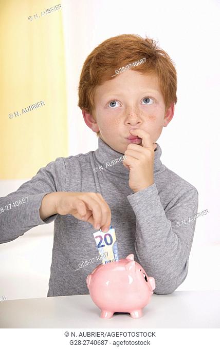Little boy with red hair, hesitating before putting a 20 euros banknote into the slot of his pink piggy bank