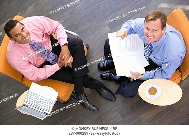 Two businessmen sitting indoors with coffee laptop and folder smiling