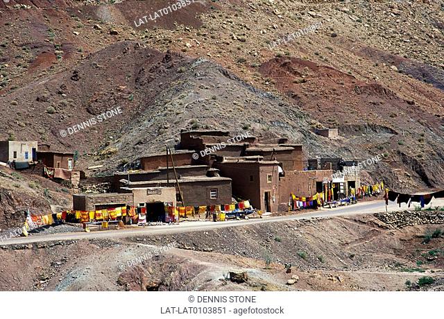 High Atlas mountains. Bare scree hillside. Mudwalled houses. Gift shop, display of fabrics, goods by road