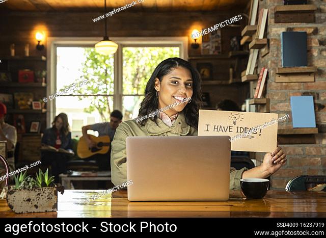Portrait of young woman sitting at table in coffee shop bookstore with laptop computer , holding hand made sign that reads ?I have ideas