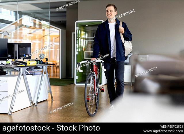 Smiling businessman with bag and bicycle walking while leaving after work from office
