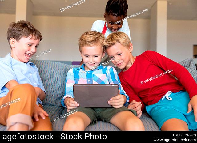 Smiling multiracial elementary schoolboys looking at digital tablet while sitting on couch in school