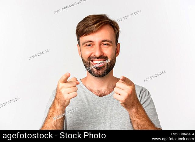Close-up of handsome adult man with beard, smiling and pointing fingers at camera, standing in gray t-shirt over white background