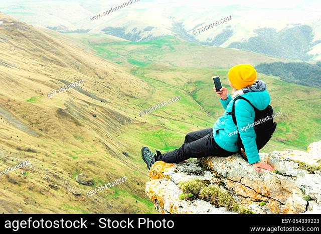 Rear view of a woman sitting on a rock in winter clothes and wearing a hat on a smartphone. Caucasus Mountains