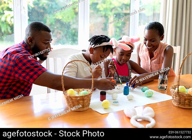 African american parents with son and daughter wearing bunny ears painting colourful eggs