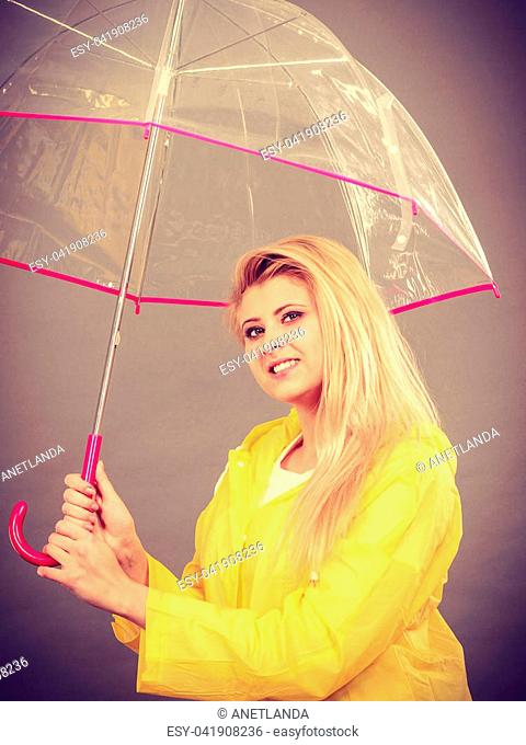 Good mood during rainy day. Happy blonde woman wearing yellow raincoat holding transparent umbrella