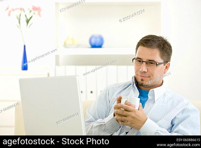 Happy casual man using laptop computer at home drinking coffee