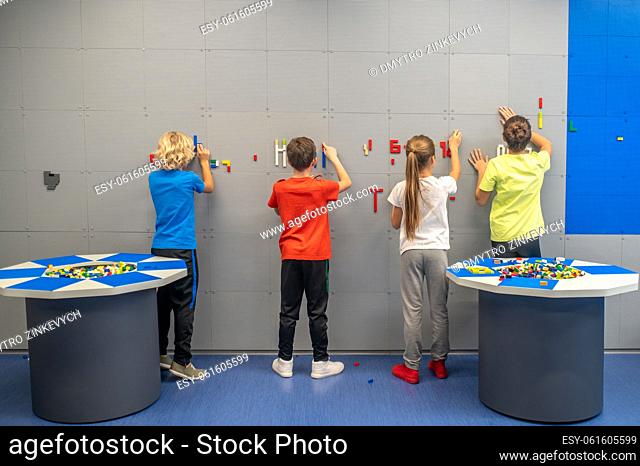 Interesting game. Back view of school children standing and attaching multi-colored building blocks to wall in playroom