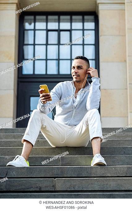 Portrait of businessman sitting on stairs listening music with earphones and smartphone in the city