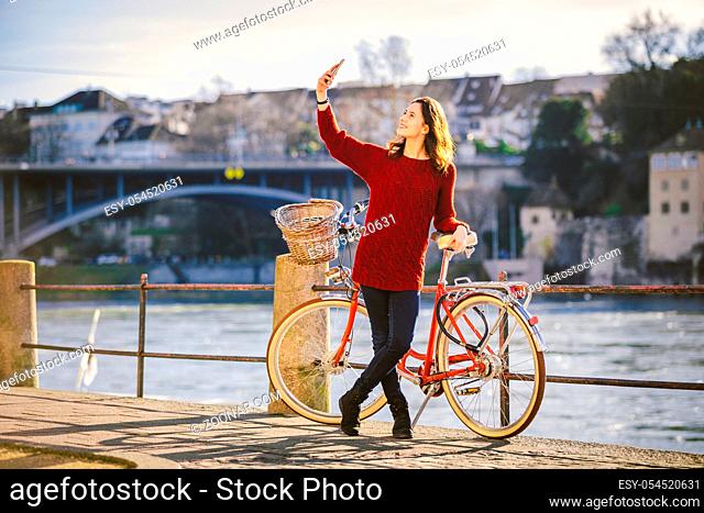 A beautiful young woman with a retro red bicycle is making a photo of herself in the old city of Europe on the River Rhine embankment in the Swiss city of Basel