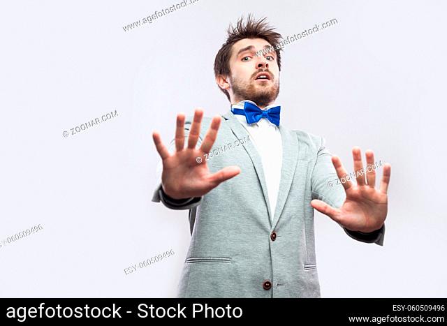 Portrait of shocked handsome bearded man in casual grey suit and blue bow tie standing with blocking hands and looking at camera scared