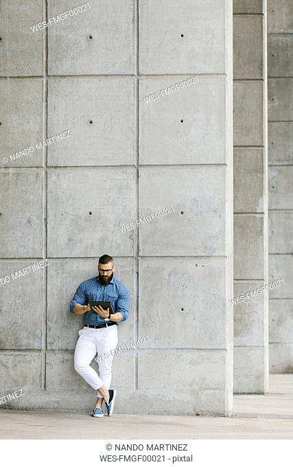 Bearded hipster businessman wearing glasses and plaid shirt leaning against wall using tablet