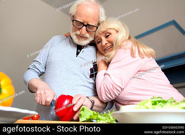 Sweet old lady hugging her husband in their kitchen. Elderly couple preparing homemade meal together cutting vegetables
