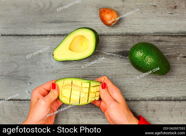 Tabletop view, young woman hands holding avocado with cuts, getting out the pulp