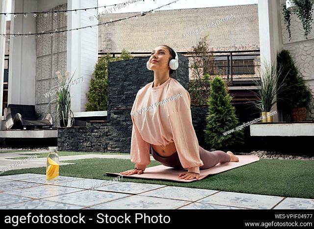 Woman listening music through wireless headphones while practicing cobra pose on terrace