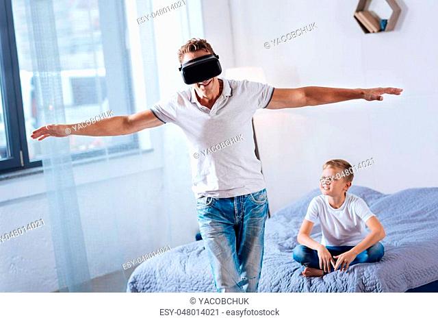 Tangible reality. Happy young man wearing a VR headset and spreading hands wide as if imitating flight while his son sitting on the bed and looking at him with...