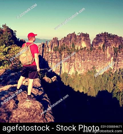 Tall tourist in red black sportswear. Sunny day in rocky mountains. Hiker with red cap, backpack and trekking poles stand on rocky view point above green valley