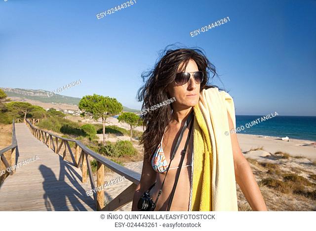 portrait of brunette summer vacation woman with sunglasses, bikini and yellow towel posing in wooden walkway, with blue sky, in Bolonia sand beach, in Tarifa