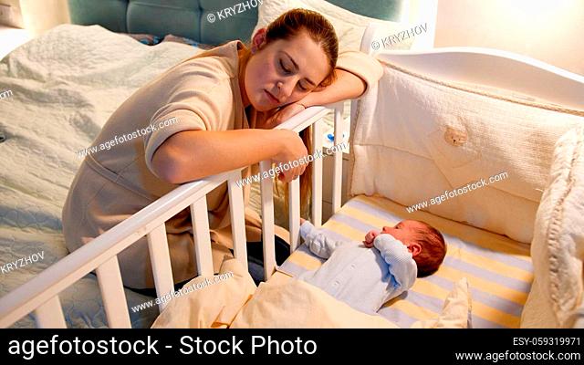 Young tired and exhausted mother fallen asleep while rocking crib of her newborn baby at night. Concept of sleepless nights and parent depression after...