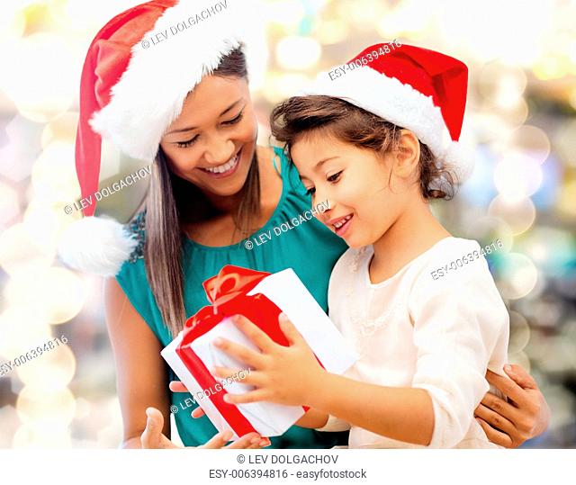 christmas, holidays, celebration, family and people concept - happy mother and little girl in santa helper hats with gift box over lights background