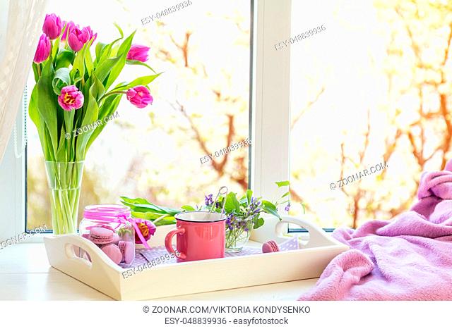 Cozy home concept. Purple fresh tulips in glass vase. Macaroons in glass jar. Cup of hot tea. White tray. Lilac blanket on the windowsill. Sunshine