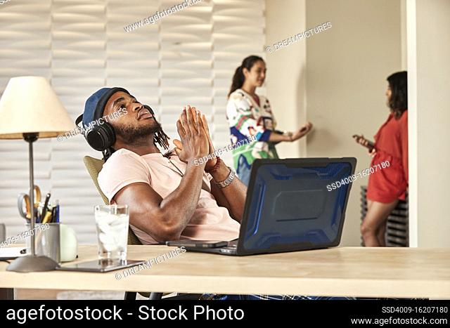 portrait young ethnic man sitting at desk with laptop computer wearing large over ear headphones, coworkers standing in background talking