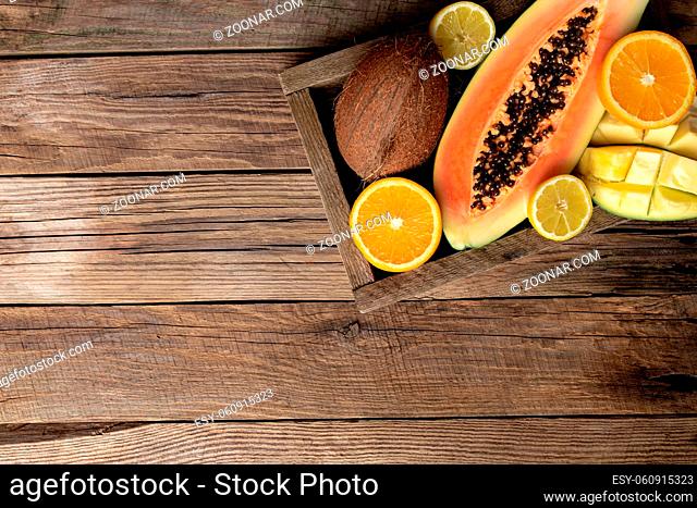 Fresh tropical fruits in a wooden delivery box on a wooden background. Papaya, orange, coconut, mango and lemon top view flat lay with copy space for some...