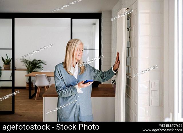 Smiling mature woman touching radiator standing at home
