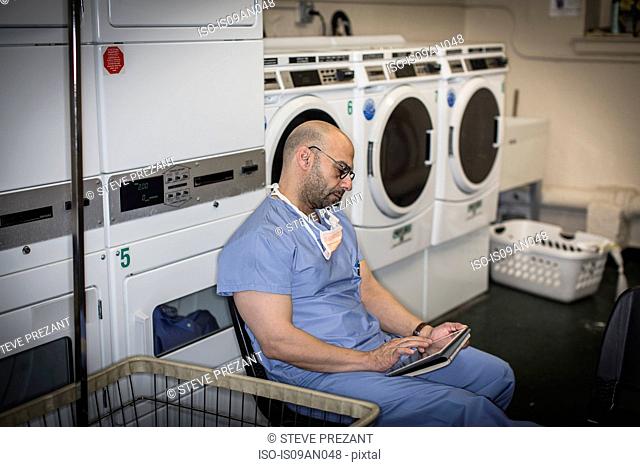 Mature man in surgical scrubs using touchscreen on digital tablet in laundry room