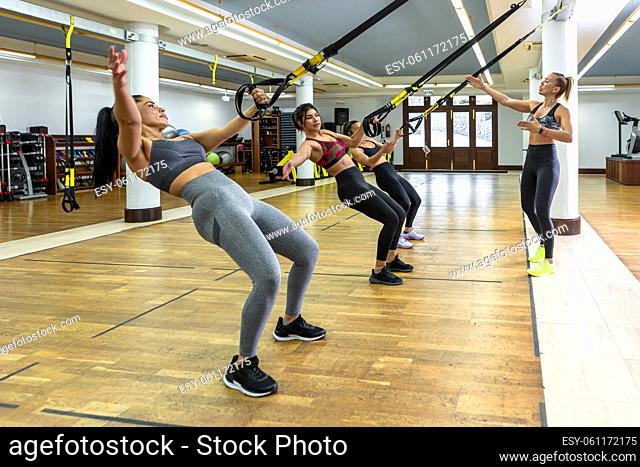 Female athletes tilting back and stretching out arms while exercising with TRX ropes near coach during suspension training in modern gym in daytime