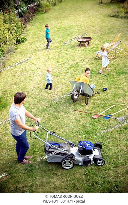 Father mowing the lawn in garden with family playing in grass