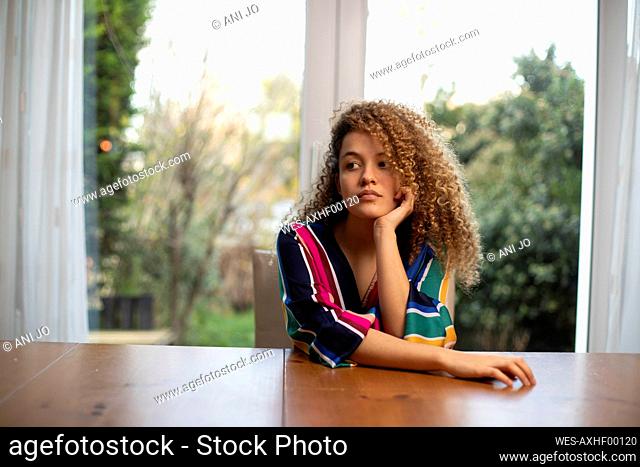 Thoughtful young woman sitting at table against window in living room