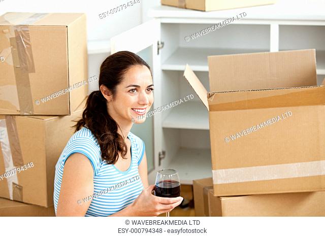 Smiling hispanic woman drinking wine between empty boxes in her new home