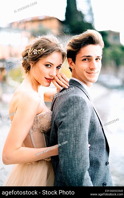 Smiling bride hugs groom from behind while standing on the promenade of Lake Como. High quality photo