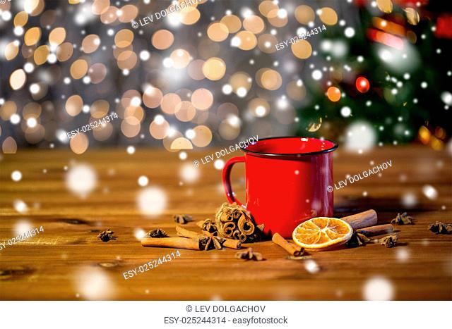 christmas, holidays, cooking and spice concept - close up of tea cup with cinnamon, anise and dried orange on wooden table over lights