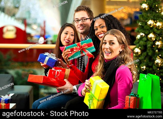 Diversity group of four people - Caucasian, black and Asian - sitting with Christmas presents and bags in a shopping mall in front of a Christmas tree with...